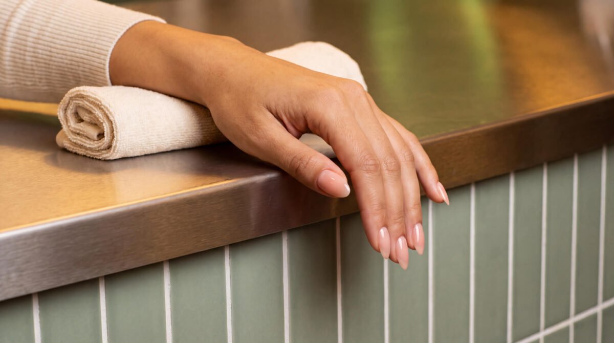 Hand with a BIAB gel manicure in soft nude, resting beside rolled salon towels